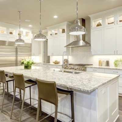 White kitchen design features large bar style kitchen island with granite countertop illuminated by modern pendant lights. Stainless steel appliances framed by white shaker cabinets . Northwest, USA